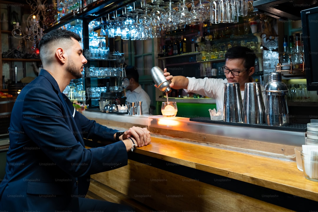 Confidence Caucasian man sitting at bar counter enjoy waiting for alcoholic drink and talking with barman in nightclub. Male mixologist bartender preparing alcohol drink in shaker with ice ball on rocks glass.